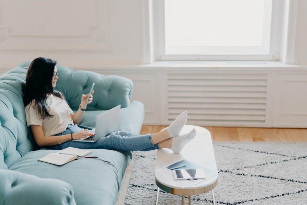 A woman is sitting on a light blue couch, using a laptop on her lap and holding a smartphone. Her legs are resting on a white coffee table next to a closed notebook and a tablet, possibly preparing for upcoming audits.