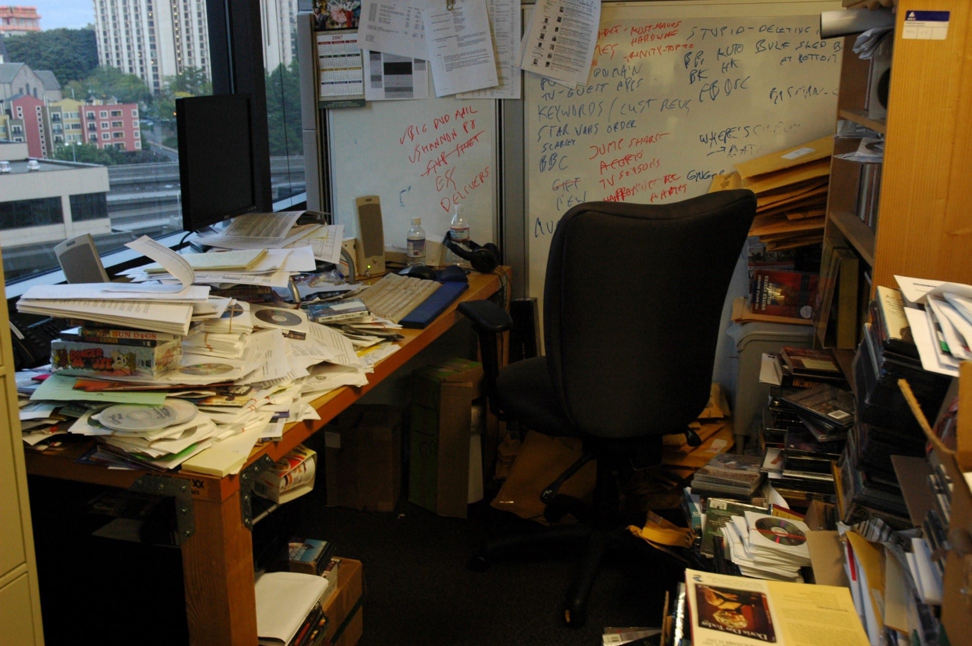A cluttered office desk with piles of papers, books, and CDs, an empty chair, and shelves filled with more documents. The background includes a whiteboard with various notes that conform to a systematic plan, and large windows showing city buildings.