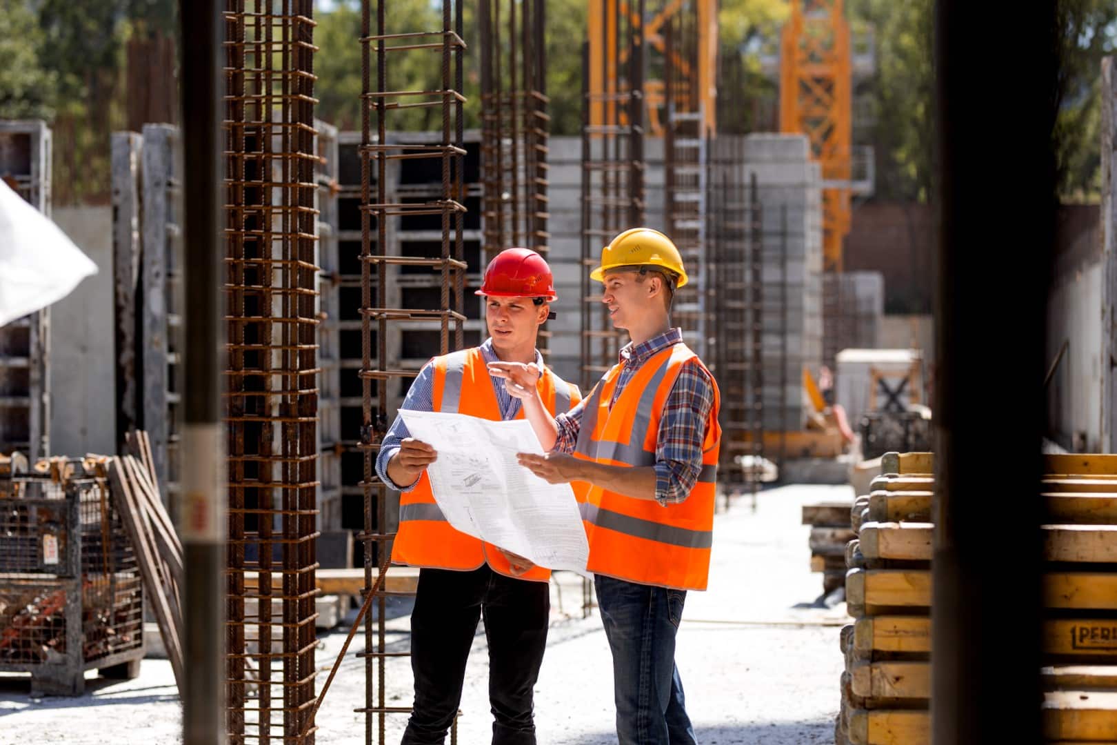 Two construction workers in safety vests and helmets review a blueprint on an active construction site with steel columns and building materials in the background, ensuring compliance with the ISO management system.