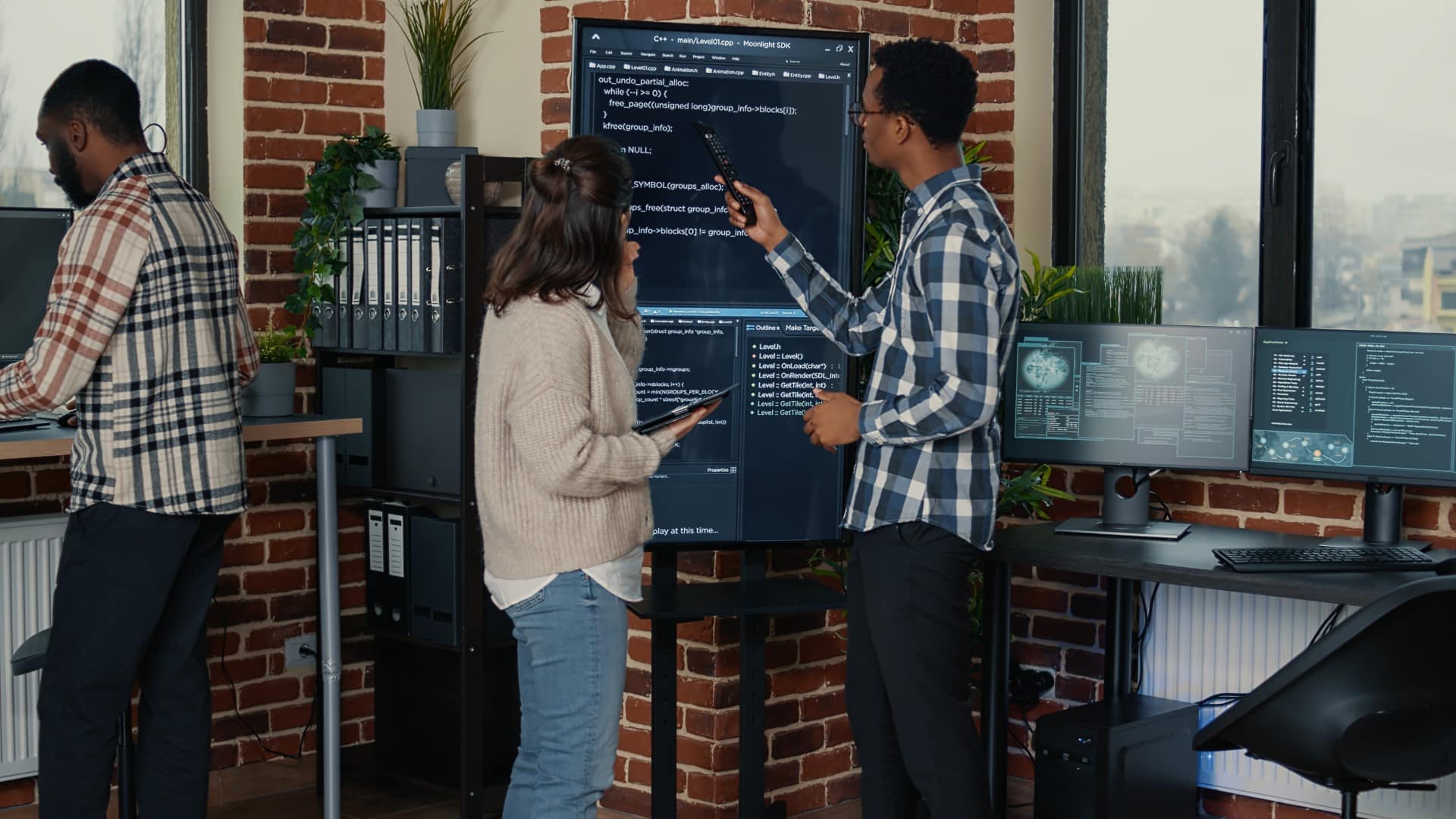 Three people are in an office with exposed brick walls. Two examine code on a large monitor, while the third person uses a computer, possibly exploring tender opportunities. Various office equipment and plants add to the productive atmosphere.