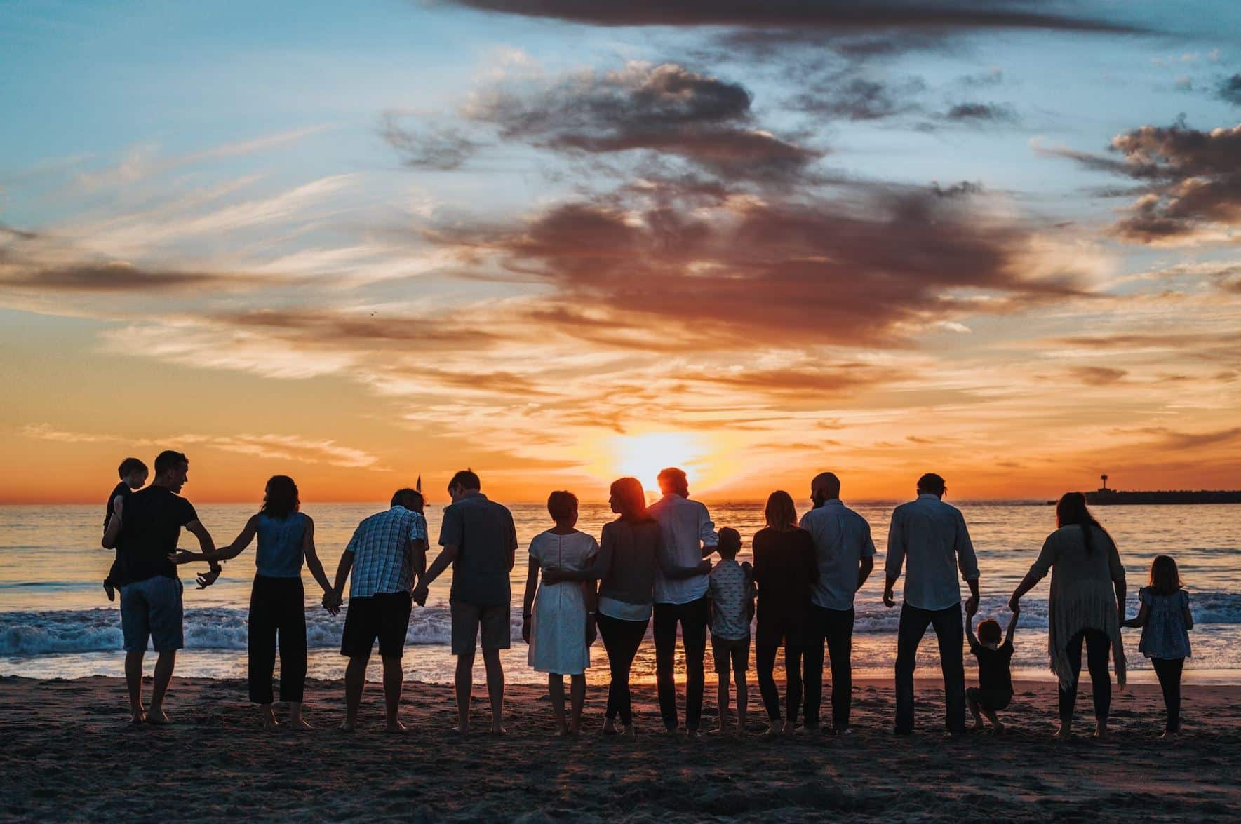 A group of people, including adults and children, stand in a line holding hands on a beach, facing a sunset over the ocean, embodying the harmony and teamwork found in an effective ISO management system.