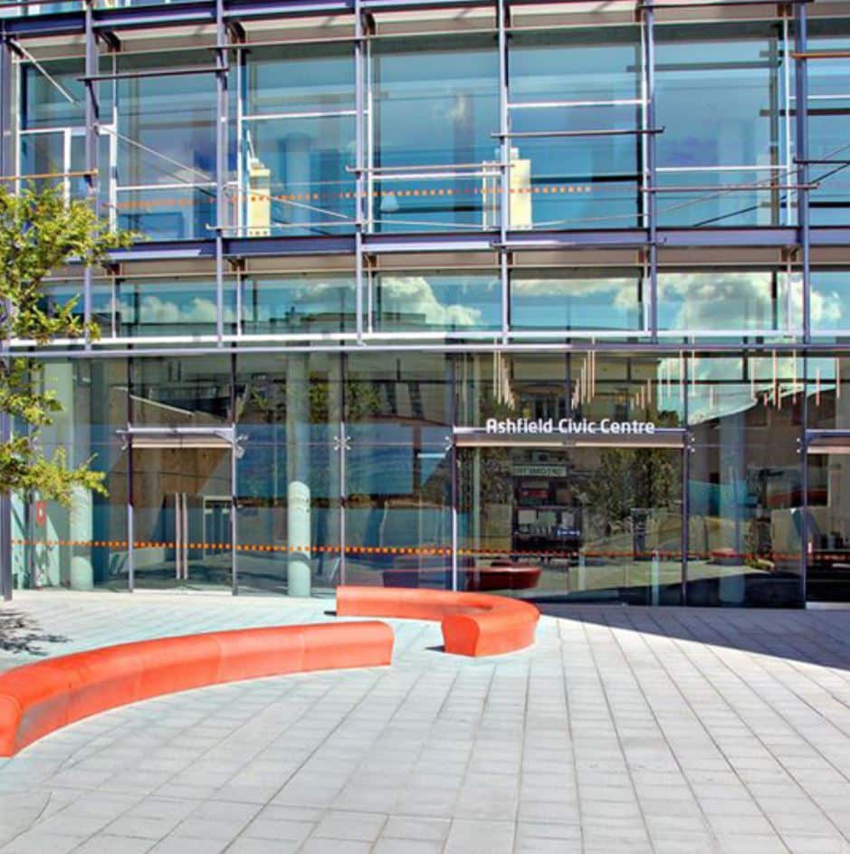 Exterior view of Ashfield Civic Centre's glass-paneled entrance with red curvilinear benches and a paved walkway in the foreground, exemplifying a commitment to process improvement through innovative design.