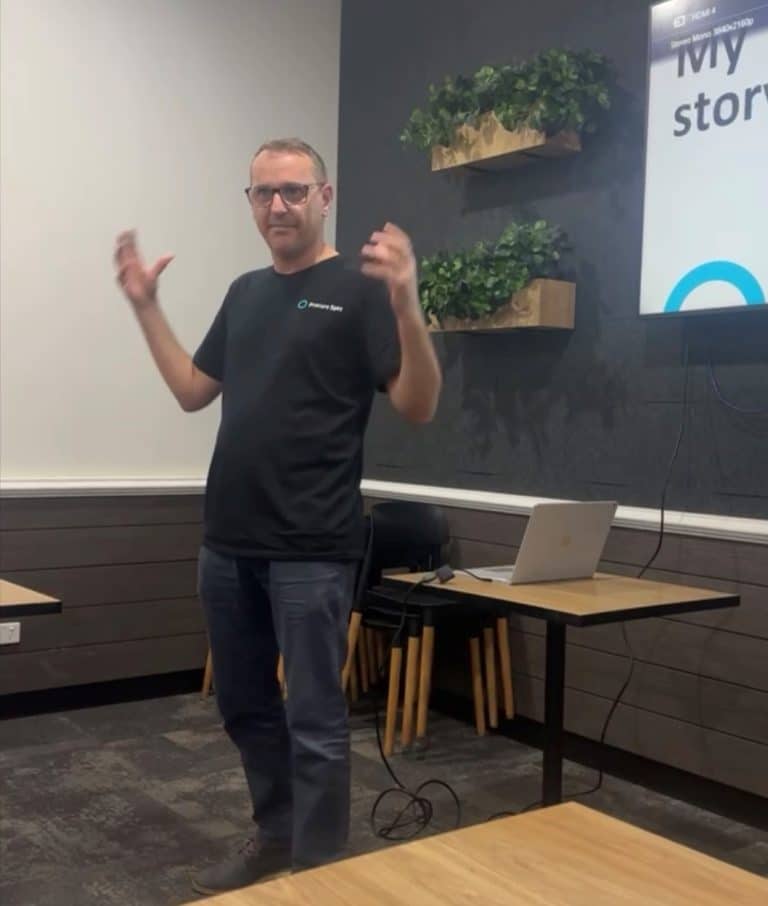 A person wearing a black t-shirt is speaking in front of a projection screen in a classroom setting. The screen, partially displaying text and connected to a laptop on the table, indicates this morning's Breakfast Talk at Penrith Chambers.