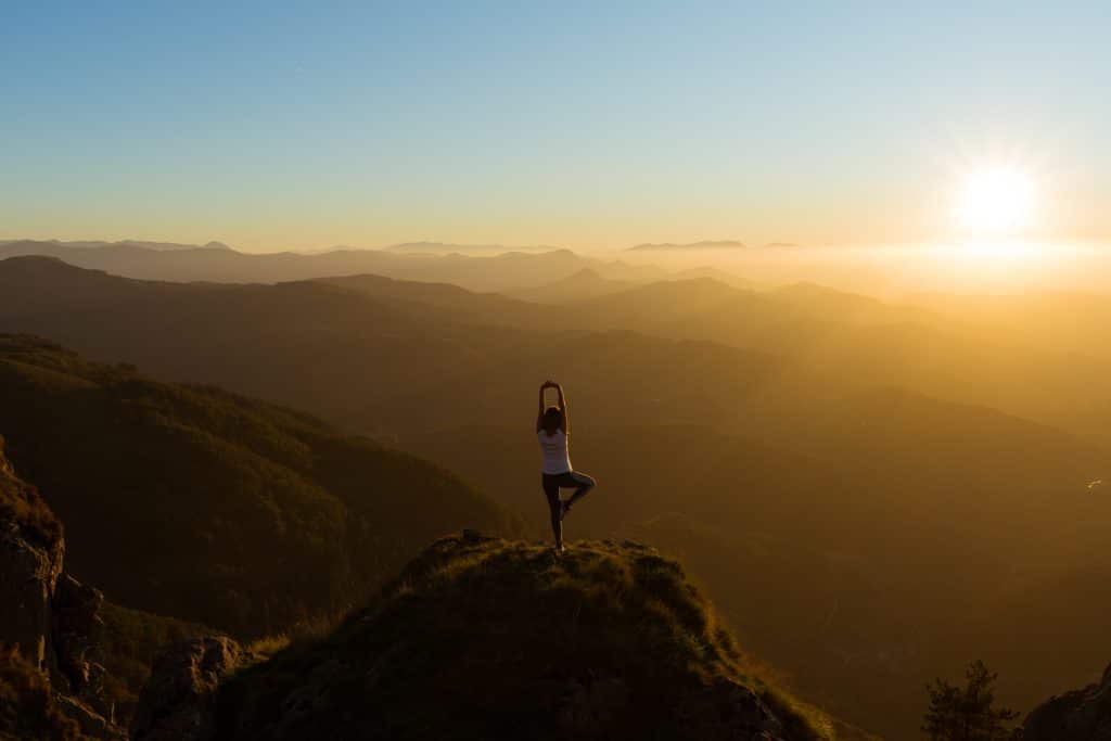 Person performing a yoga pose on a mountain peak at sunset, overlooking a vast landscape of hills and valleys, embodying the tranquility needed for scaling your small business successfully.