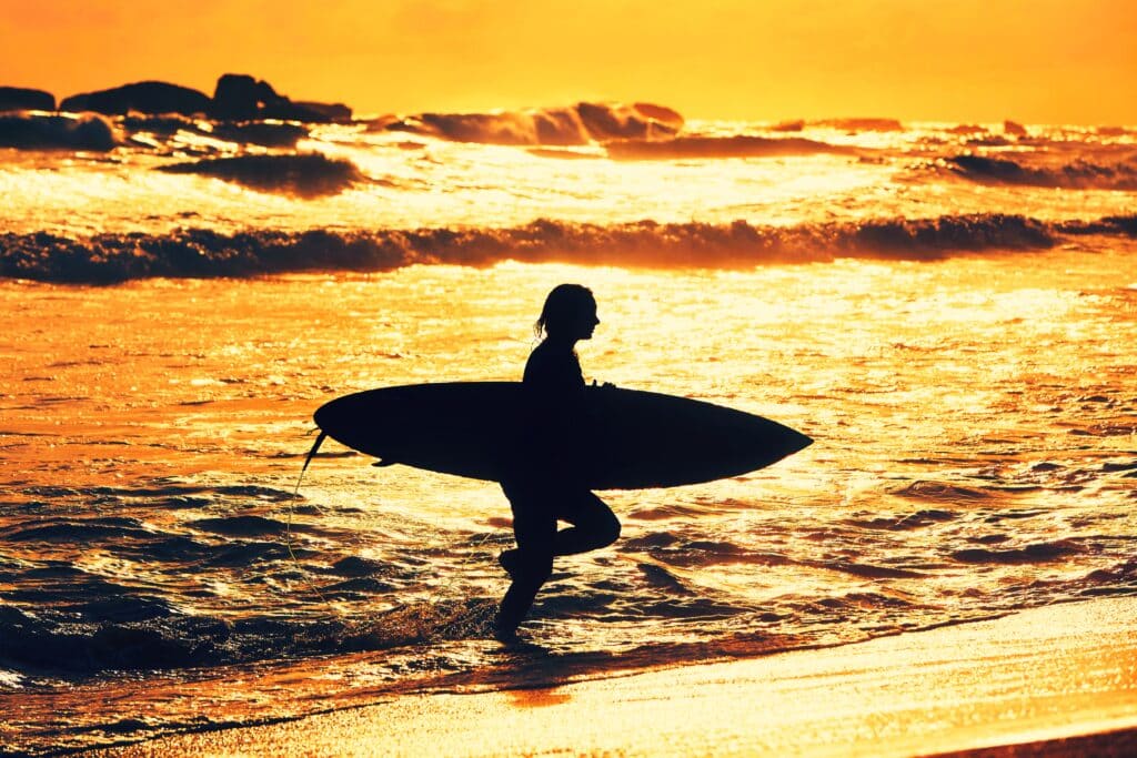 A person holding a surfboard walks through shallow ocean water at sunset, with waves crashing in the background, reflecting on the parallels between mastering surfing and mitigating cybersecurity risks.