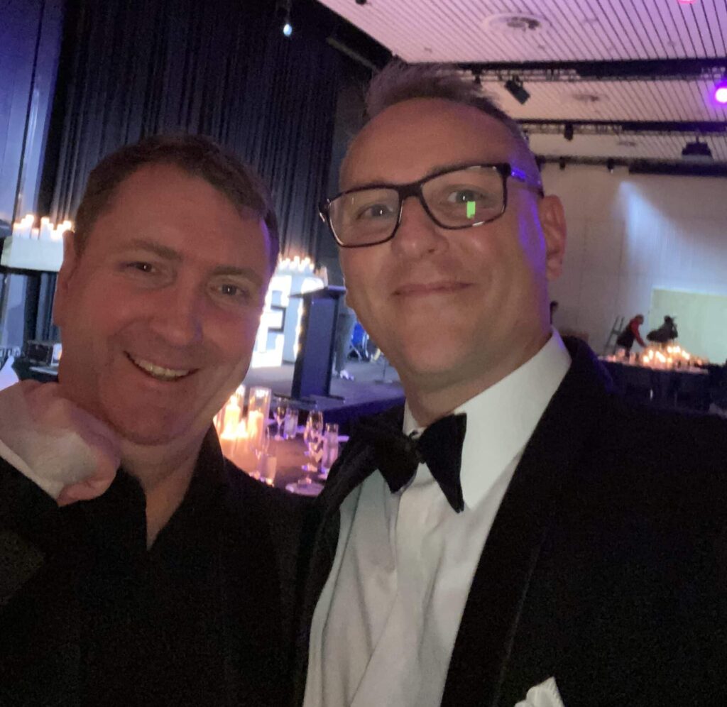387d63e0 e314 42d4 9e31 411f495627fe scaled e1724808582901 Entrepreneurs' Organisation's (EO) Board Handover & Gala Night - 1 Two men in formal attire pose for a selfie at an indoor event hosted by the Entrepreneurs' Organisation, with candlelit tables and dark drapery in the background.