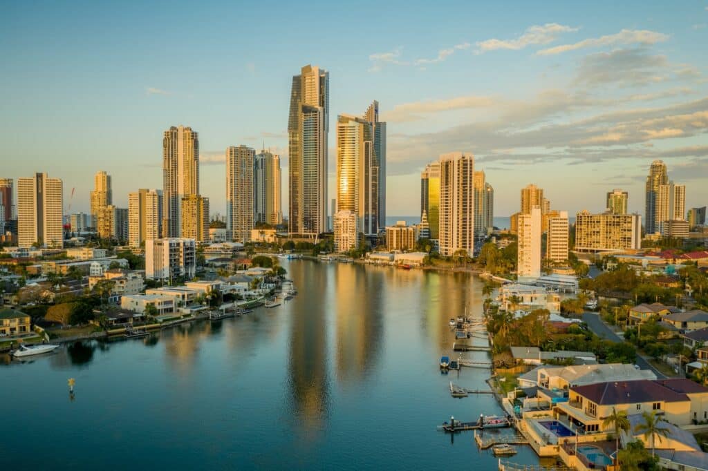 Aerial view of a city skyline with numerous high-rise buildings reflecting in a calm river during sunset, evoking an Australian business hub. Residential houses line the riverbanks in the foreground, adding a quaint charm to this bustling procurement context.