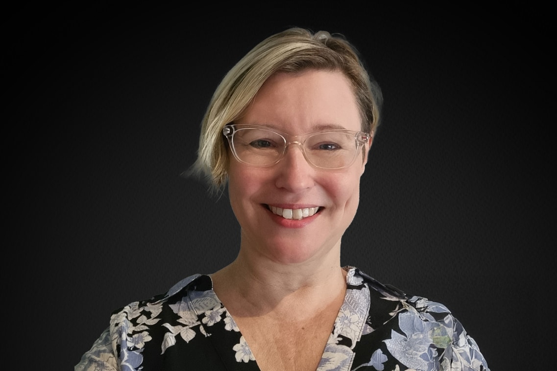 2ca6c1a5 01d5 4704 ae06 8fa4e8ce389a About Us - 9 A woman with short blonde hair and clear glasses smiles at the camera, wearing a floral-patterned top against a plain dark background.